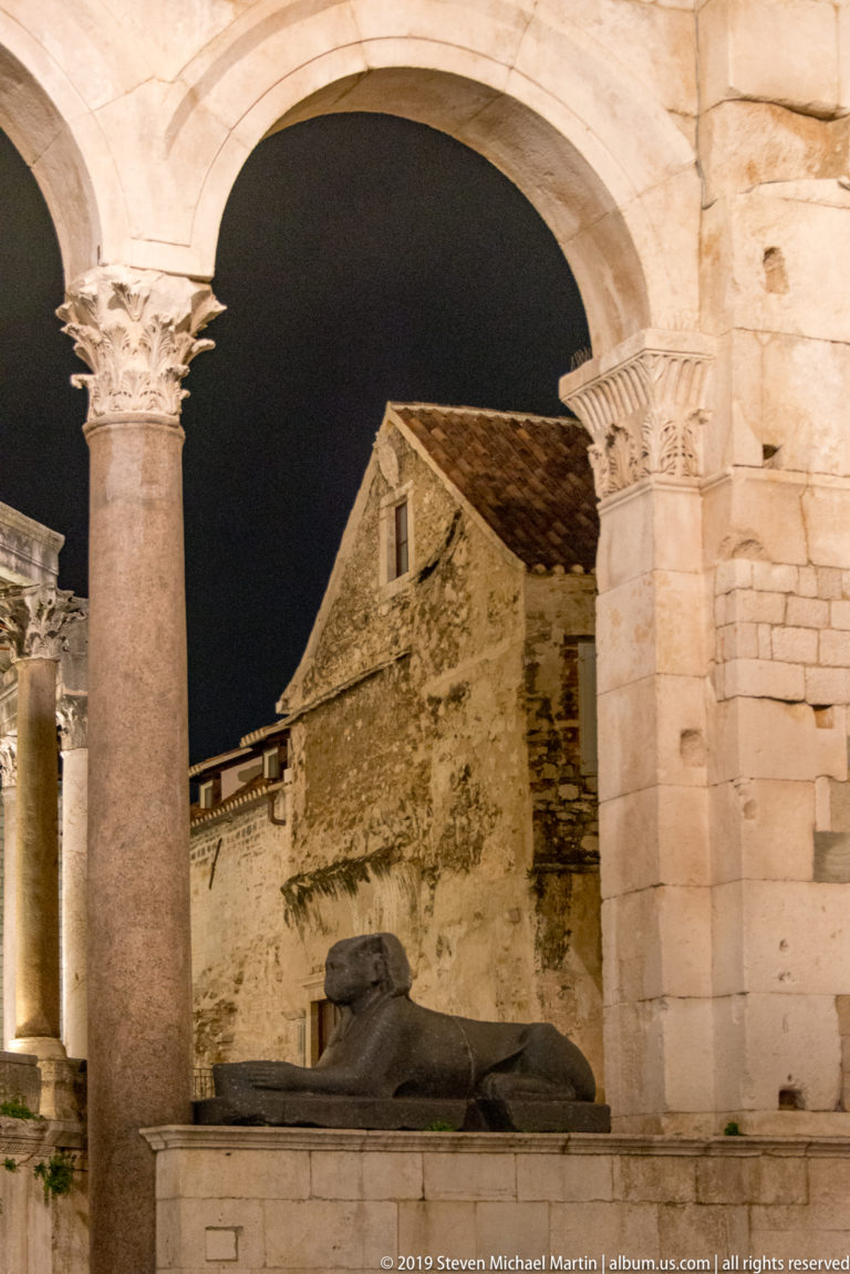 Stone sphinx rests beneath illuminated columns and arches of the Peristyle, with a rough masonry building behind it at night.