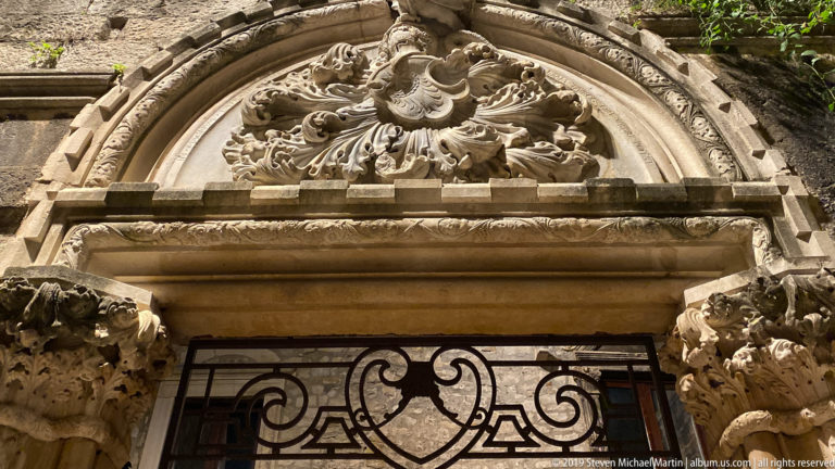 Ornate historic stone doorway with carved rounded pediment, scrollwork, columns, and a black wrought-iron grille.