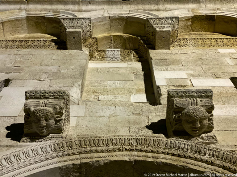Two carved stone lion heads project from a pale masonry façade above an ornate horizontal band at night.