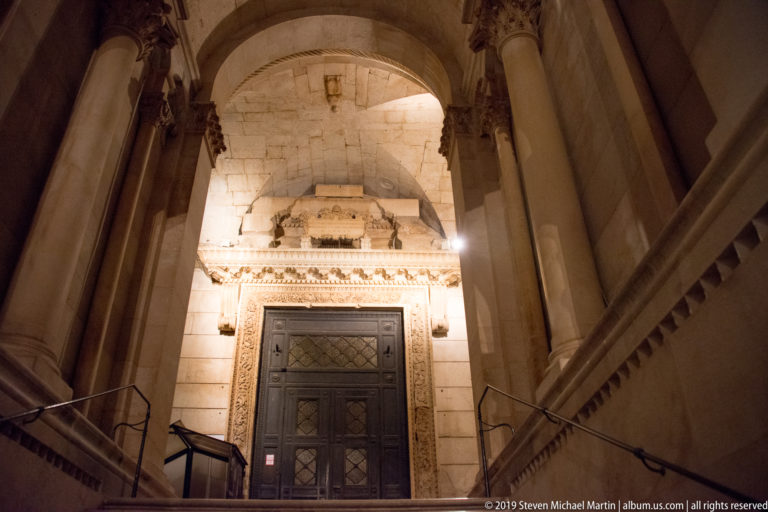 Stone steps rise to the dark wooden entrance of Saint Domnius Cathedral beneath an illuminated arch and flanking columns.