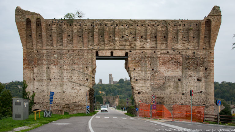 Road passing through a large opening in the weathered brick Ponte Visconteo, with a distant castle tower visible beyond.