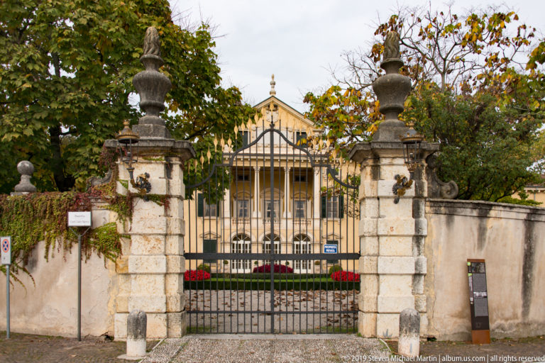 Wrought-iron gate between urn-topped stone piers, with a yellow historic villa visible through autumn trees beyond.