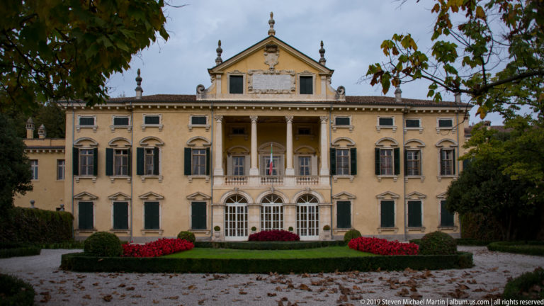 Yellow Villa Maffei façade with central portico, green shutters, formal hedges, red flowers, and fallen autumn leaves.