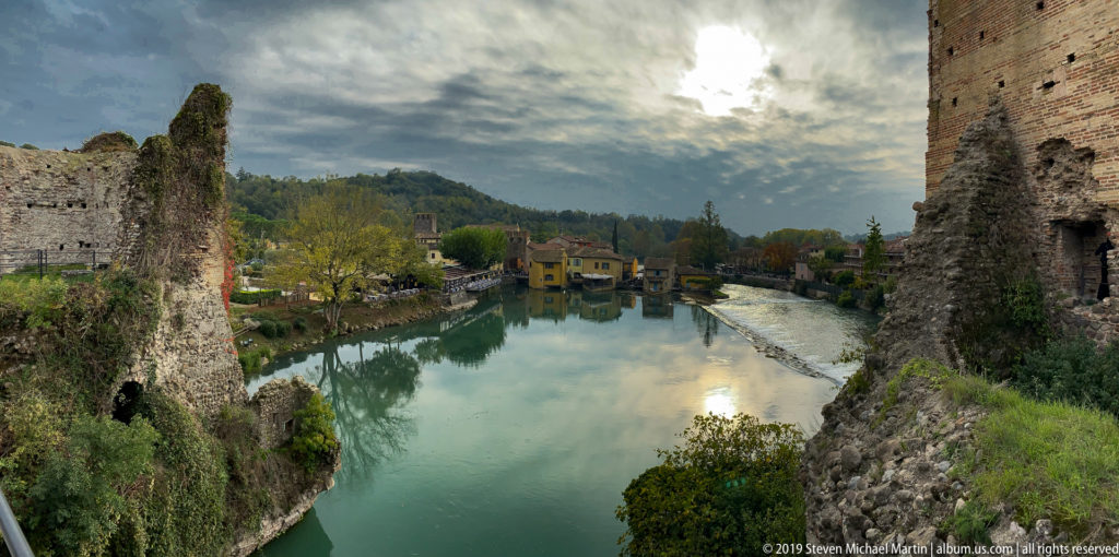 Mincio River flowing between old masonry fortifications toward Borghetto houses, with a low weir at right.