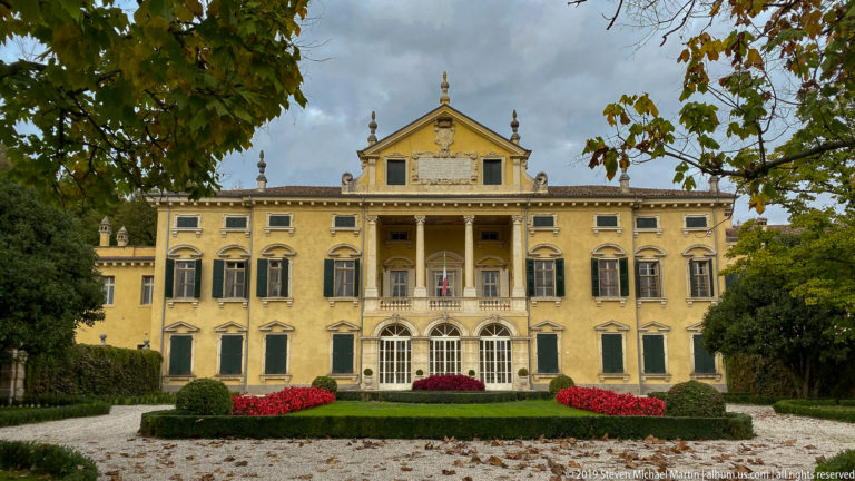 Closer frontal view of a yellow historic villa with central portico, green shutters, roof ornaments, hedges, and red flowers.