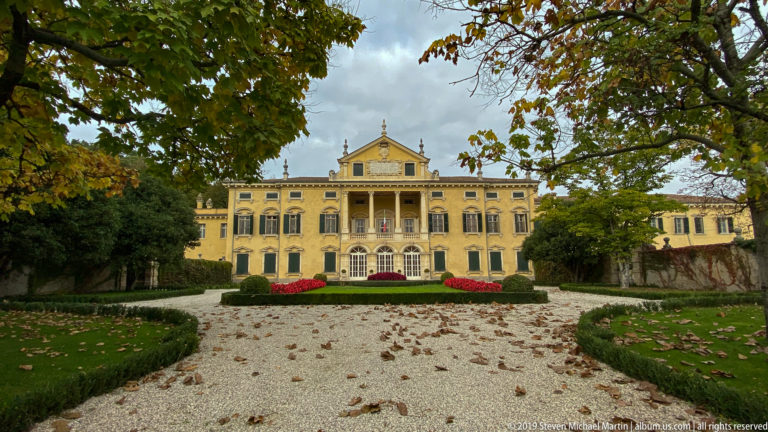 Yellow stucco façade of Villa Maffei beyond a gravel garden path, clipped hedges, flower beds, and autumn trees.