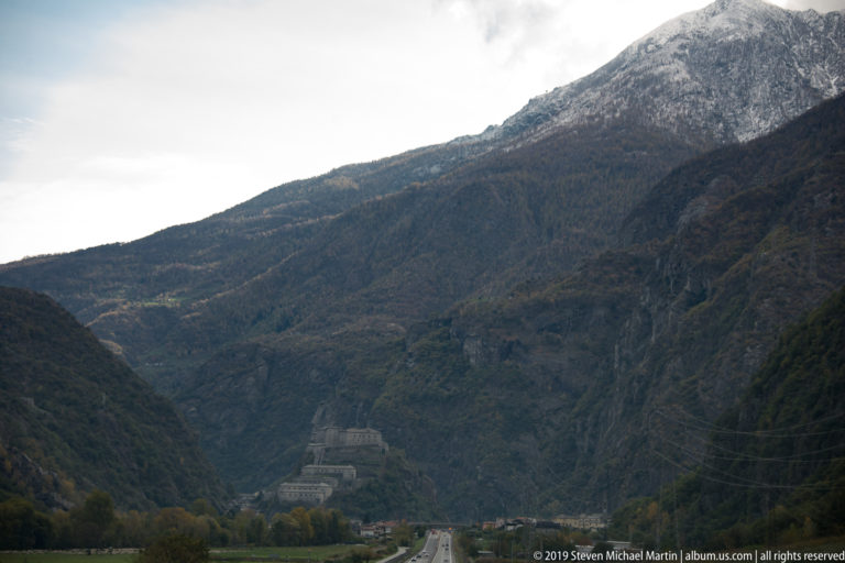 Road running through a narrow Alpine valley beneath steep wooded and rocky slopes in overcast weather.
