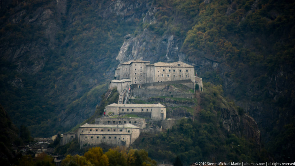 Distant view of Forte di Bard beneath rocky, forested Alpine slopes, with lower buildings descending toward the valley.
