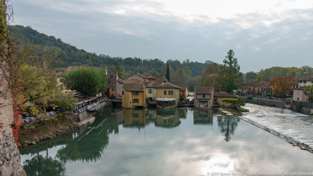 Wide view over Borghetto and the reflective Mincio River, framed by autumn trees and old masonry at left.