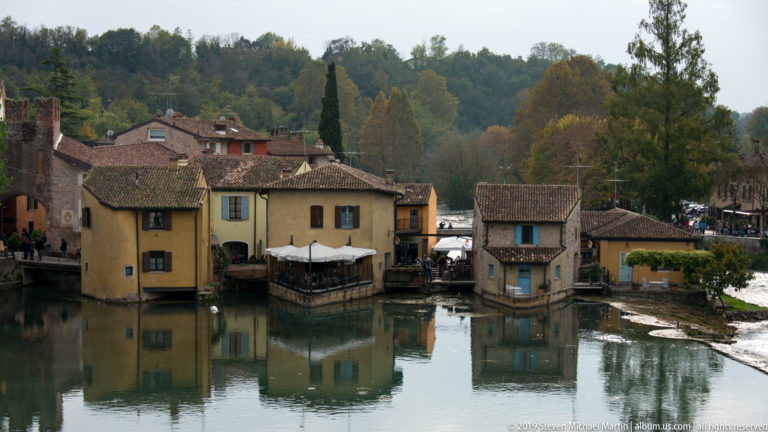 Elevated view of Borghetto houses reflected in the Mincio River, with a low weir curving along the right side.