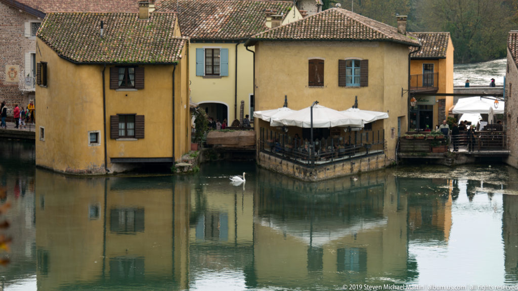 Yellow riverside buildings and café terrace reflected in the Mincio, with a white swan swimming in front.