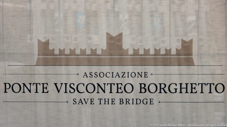 Sign reading “Associazione Ponte Visconteo Borghetto — Save the Bridge” beneath a stylized silhouette of the fortified bridge.