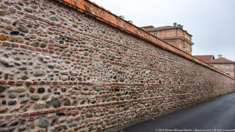 Long historic wall of rounded stones and brick courses extending beside a narrow road, with palace buildings beyond.