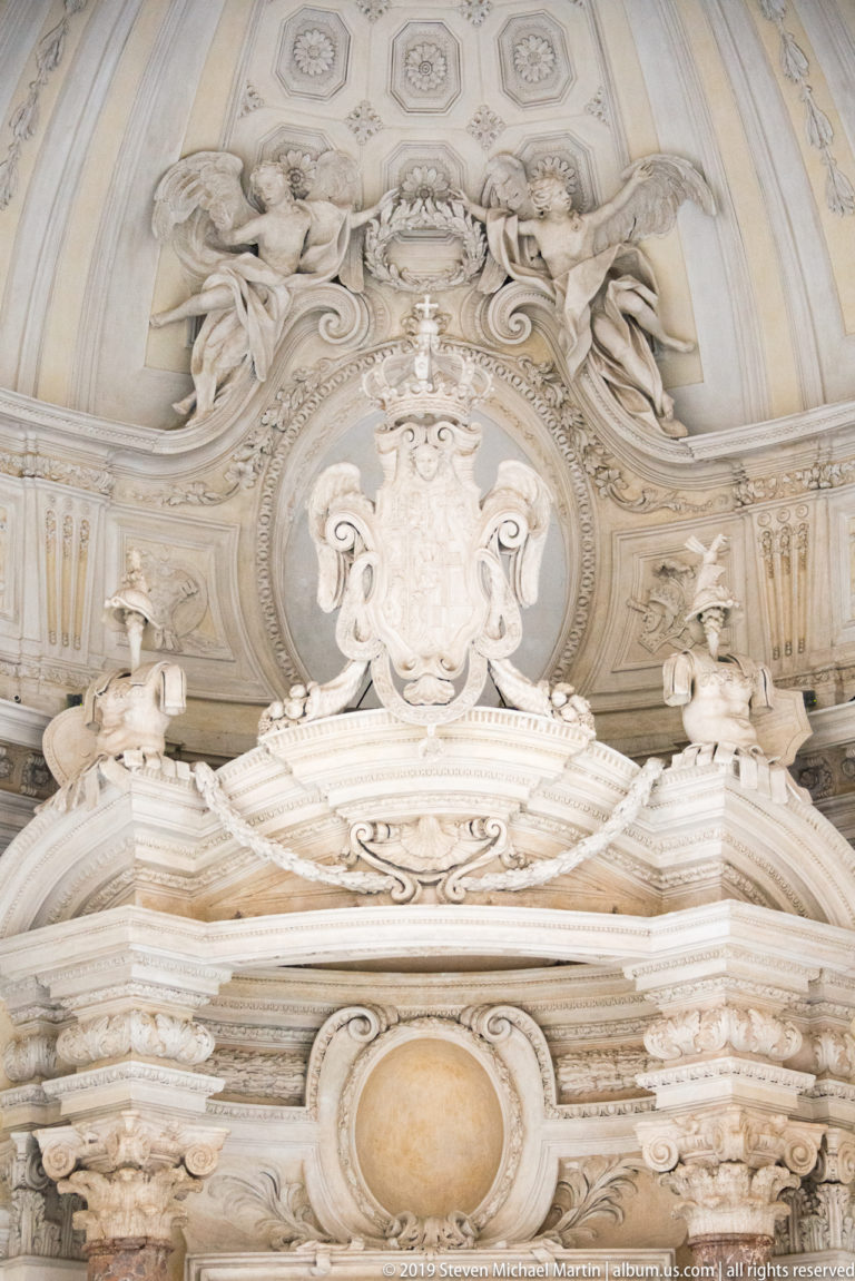 Ornate pale stucco overdoor with central cartouche, reclining figures, layered cornices, and scrolling decoration.