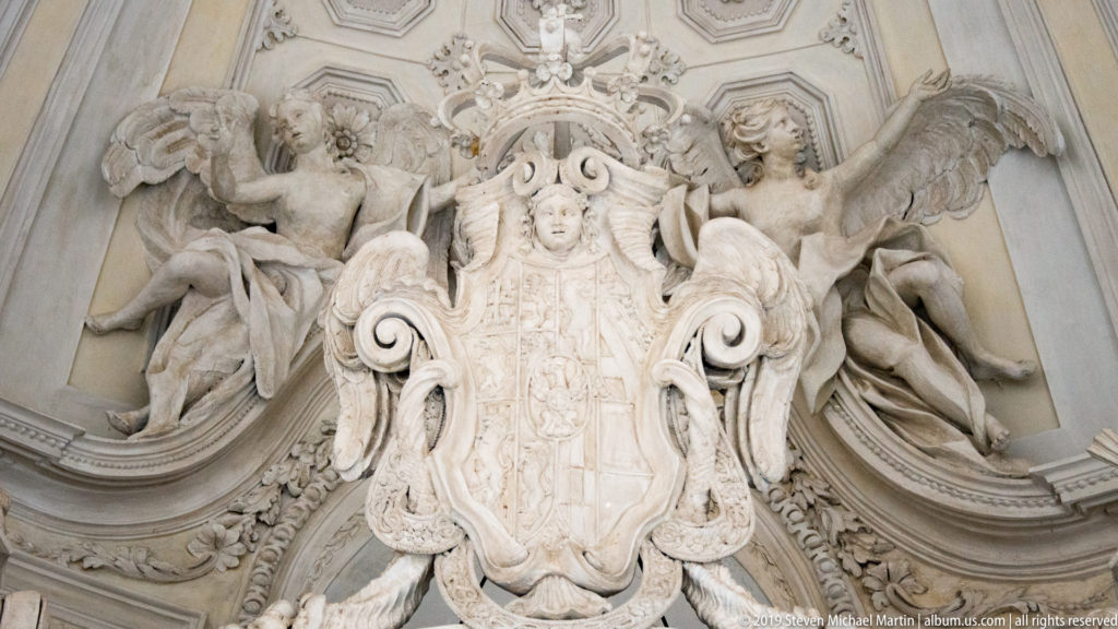 Large pale stucco cartouche with shield, scrolling ornament, and two winged supporting figures in high relief.