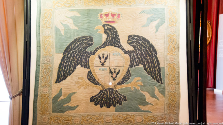 Embroidered textile with a black double-headed eagle, crowned multi-field heraldic shield, and decorative border.