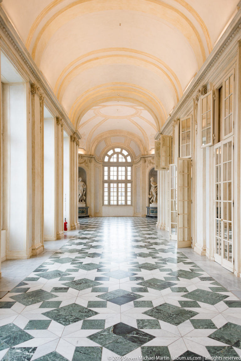 Wide vaulted gallery with open windows and doors, green-and-white geometric floor, and statues at the far end.