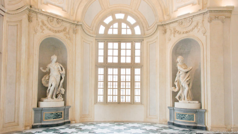 Two white classical statues on green-veined pedestals flanking a tall arched window above a geometric stone floor.