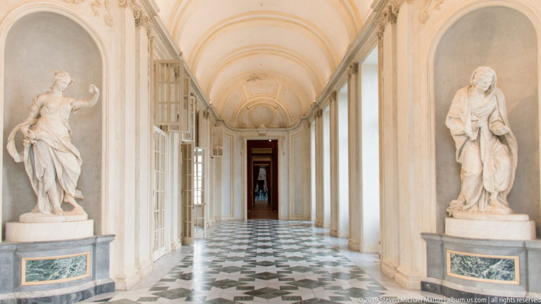 Long vaulted palace corridor with white statues on pedestals, tall windows, and a geometric black-and-white marble floor.