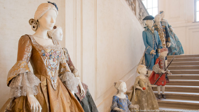 Mannequins in embroidered court gowns and blue coats arranged on a staircase inside La Venaria Reale.