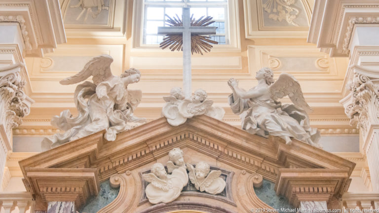 Large cross above a triangular pediment crowded with white angel and cherub sculptures inside the Church of Sant'Uberto.
