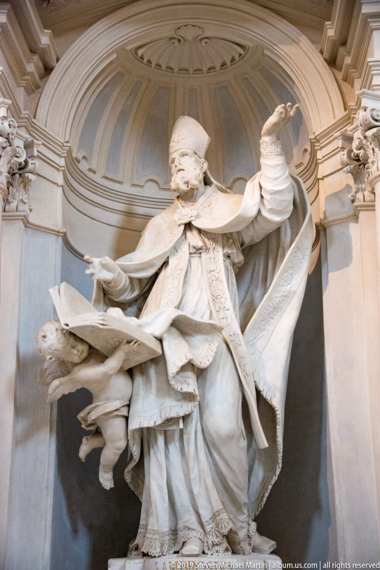 Large white statue of a robed cleric or saint raising one arm within an ornate architectural niche.