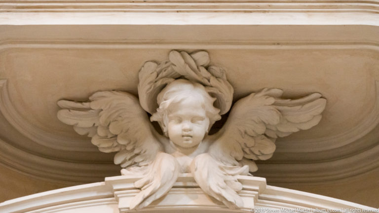 Close view of a pale stucco winged cherub head projecting above an interior architectural molding.