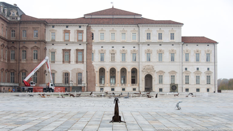 Wide courtyard façade of La Venaria Reale with brick and pale stucco sections, animal sculptures, and a maintenance lift.