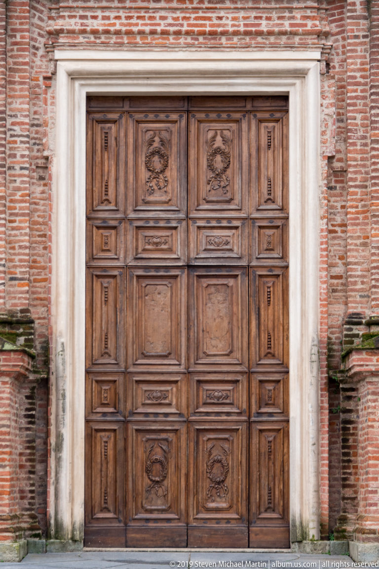 Large paneled wooden door with carved medallions in a pale stone surround set into a red-brick wall.