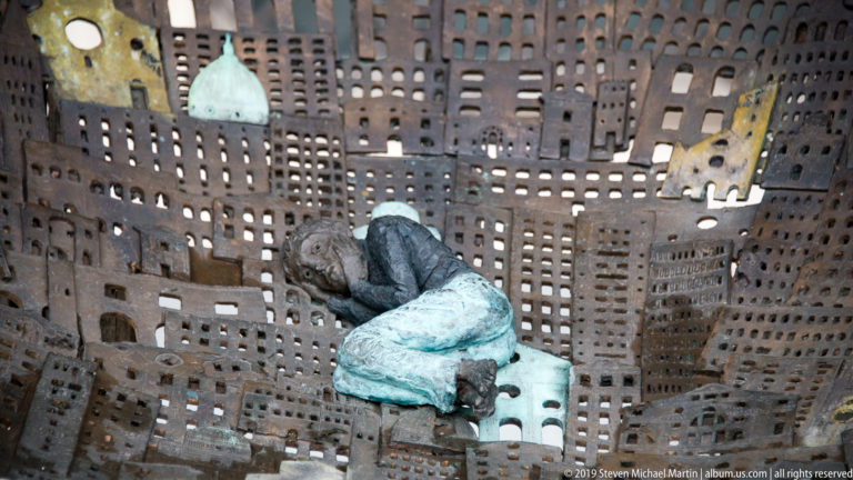 Small seated figure in muted clothing positioned inside a perforated metal structure patterned like dense city buildings.