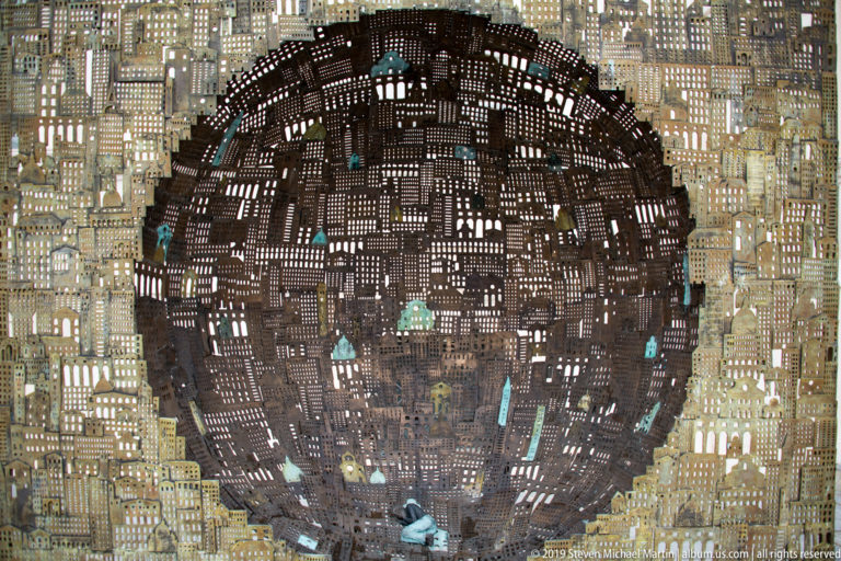 Large circular perforated metal sculpture resembling a dense miniature city, with small colored figures embedded within it.