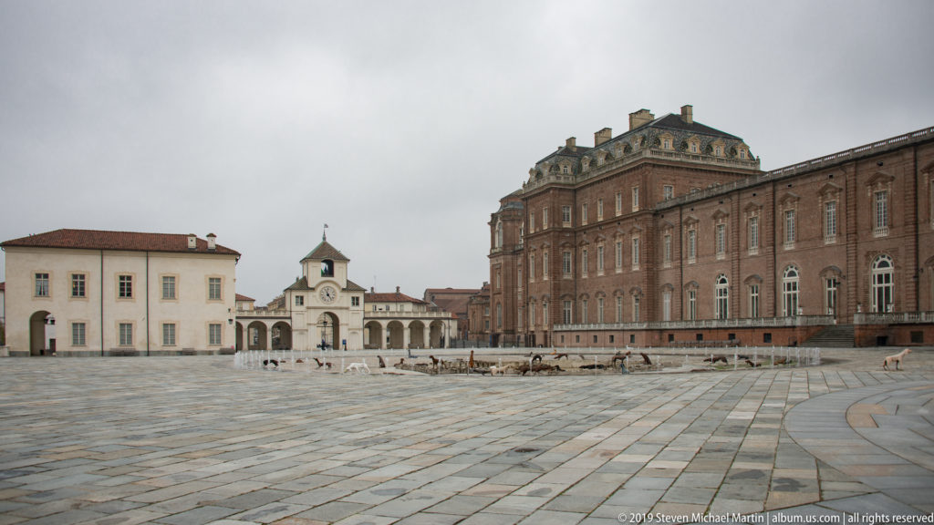 Broad paved courtyard between a tall brick palace wing and lower pale buildings with a small central tower.