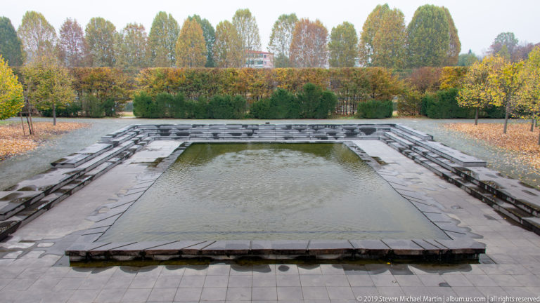 Rectangular sunken water basin bordered by stepped dark stone platforms and autumn trees.
