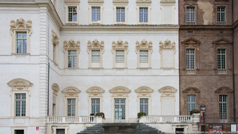Ornate white palace façade with sculpted window surrounds meeting a darker exposed-brick elevation at right.