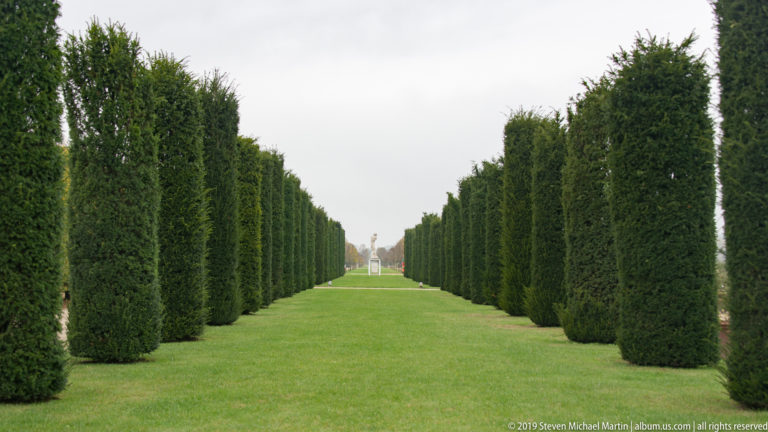 Long grass avenue enclosed by tall clipped hedges, converging toward a distant statue.