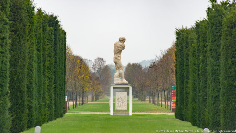 Pale figurative statue on a pedestal centered at the end of a long avenue between tall clipped hedges.