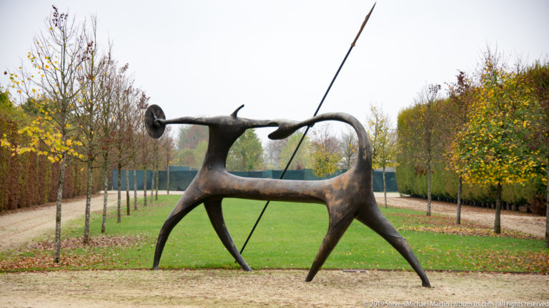 Stylized bronze animal sculpture on long legs with a tall diagonal pole, set along a tree-lined garden axis.