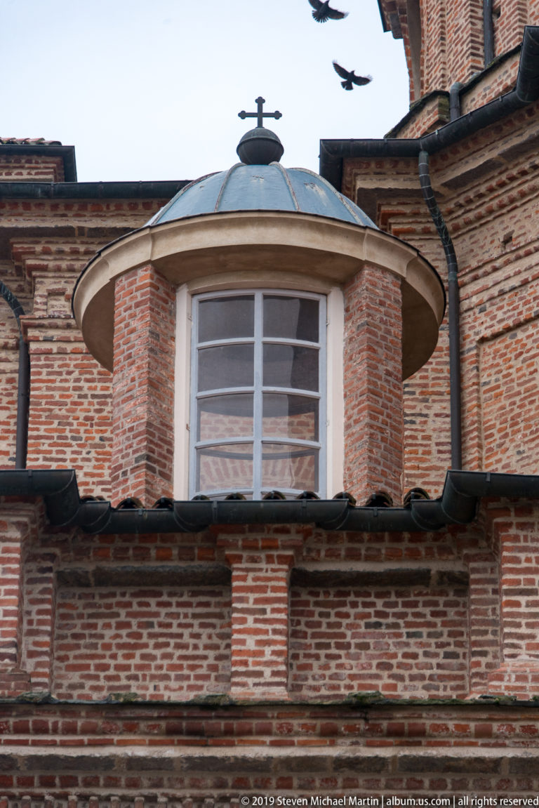 Curved projecting window with white glazing, shallow metal roof, and finial set into a red-brick palace roofline.
