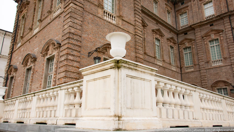 White stone balustrade and large urn beside a richly detailed brick palace façade with tall windows.