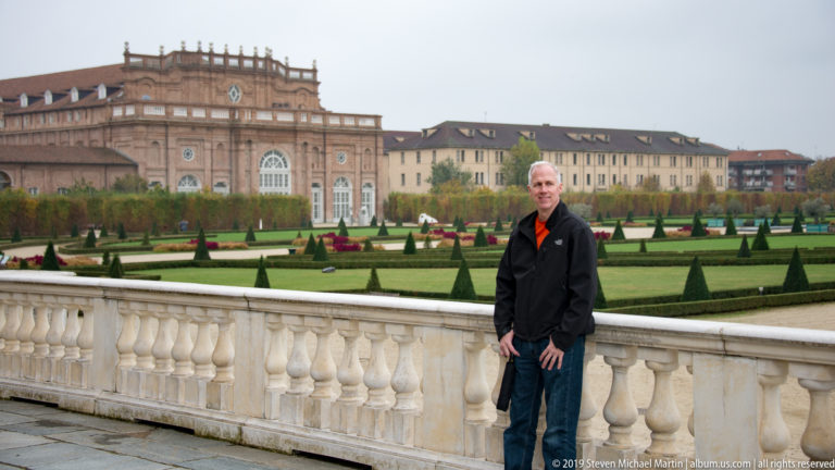 Man standing beside a pale stone balustrade overlooking clipped formal gardens and palace buildings.