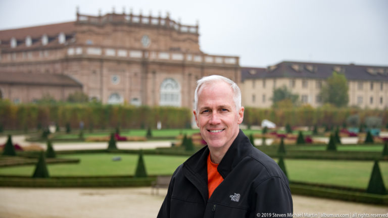 Smiling man in a black jacket standing before geometric hedges, topiary, and palace buildings at La Venaria Reale.