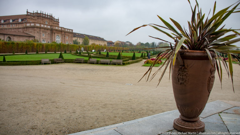 Large brown ornamental urn with spiky plant on a terrace above clipped formal gardens and palace buildings.