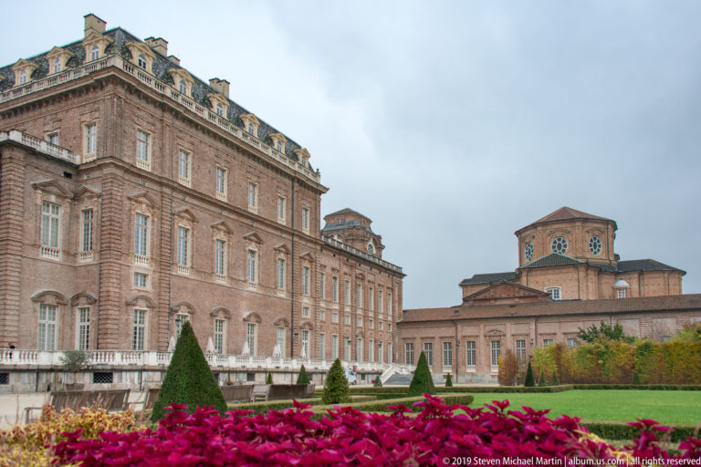Brick palace wing beside a lower range with square clock tower, seen beyond topiary and red garden foliage.