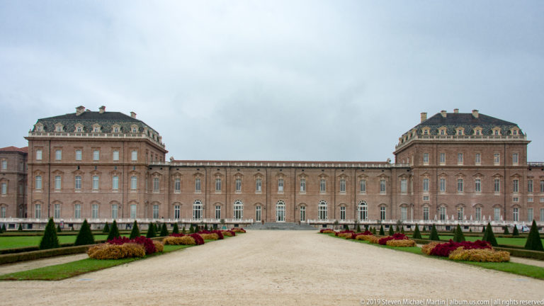 Wide gravel axis lined with topiary and low beds leading straight toward La Venaria Reale’s long brick façade.