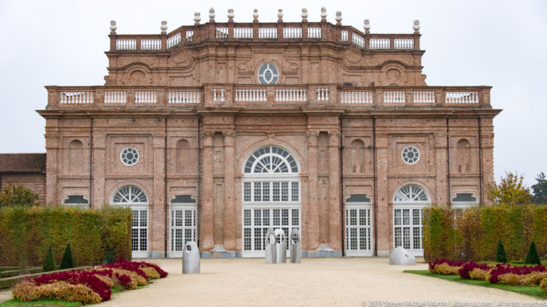 Ornate brick façade with tall central glazed doorway, columns, arched windows, balustrades, hedges, and flower beds.
