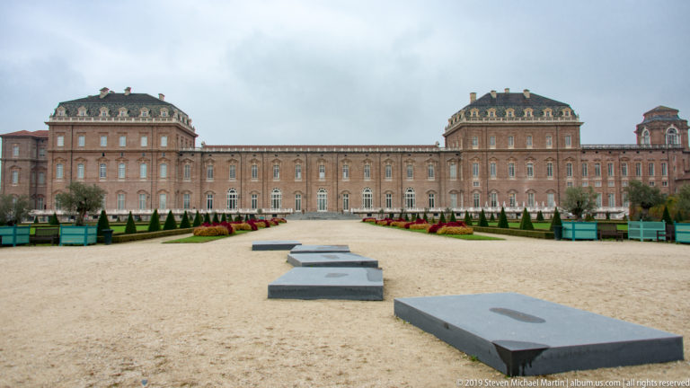 Long symmetrical brick palace façade facing a broad gravel parterre with clipped hedges, topiary, and red flower beds.