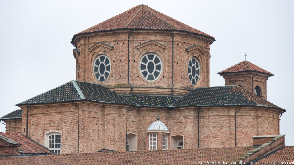 Polygonal brick church structure with large oval windows rising above tiled roofs within the La Venaria Reale complex.
