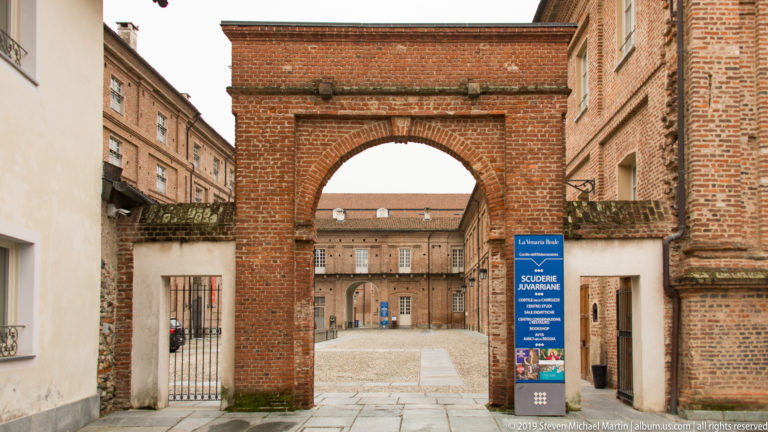 Brick arched gateway framing an inner courtyard and palace building at La Venaria Reale, with an exhibition sign beside it.