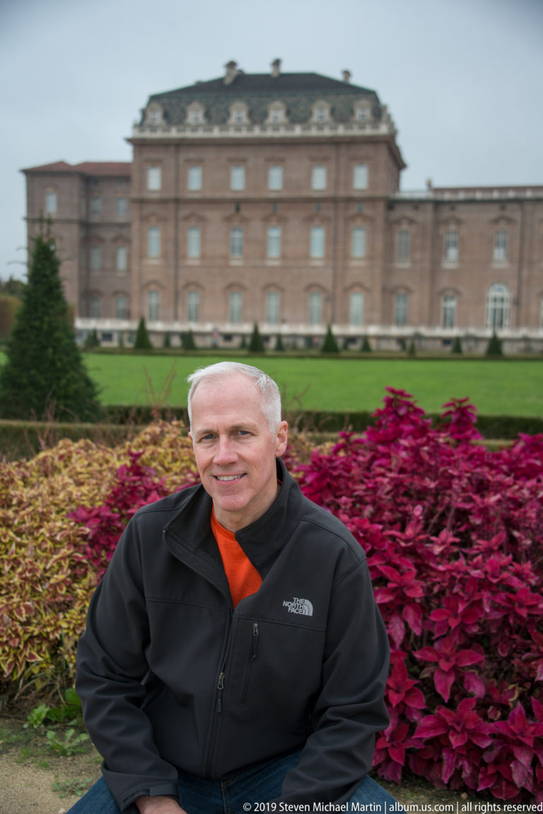 Smiling man in a black jacket seated before burgundy garden foliage, with the palace softly focused behind him.
