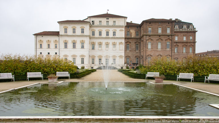 Circular fountain basin with a small central jet, benches, and the pale and brick palace façade behind.